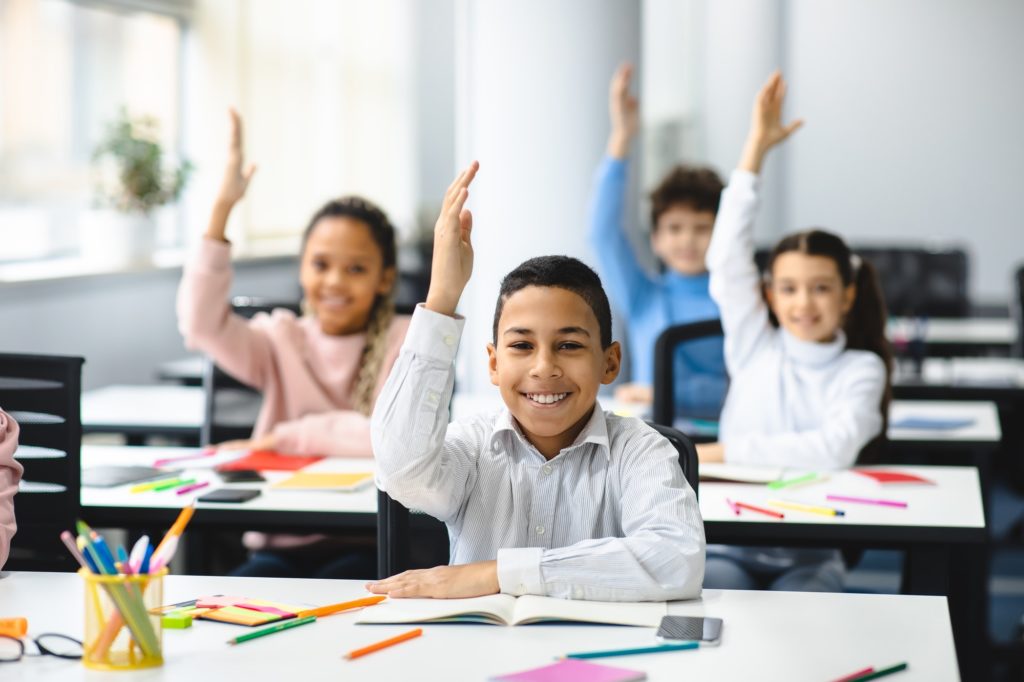 Diverse small schoolkids raising hands at classroom