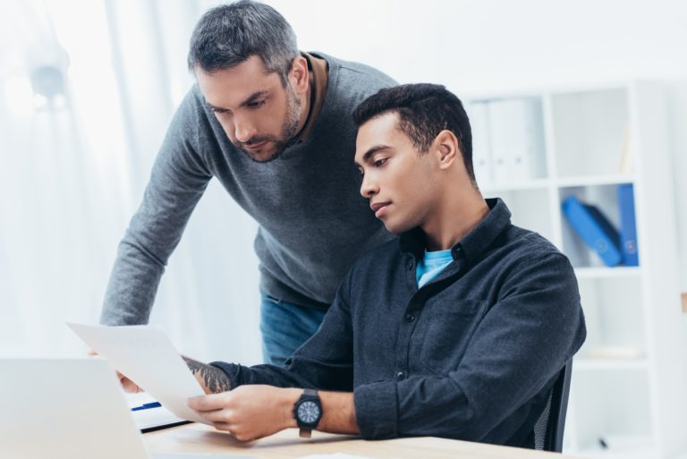 bearded mentor looking at young colleague working with papers in office